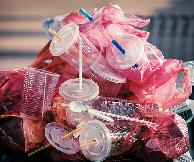 Heap of plastic cups, straws, and spoons with pink plastic bags, highlighting plastic waste.