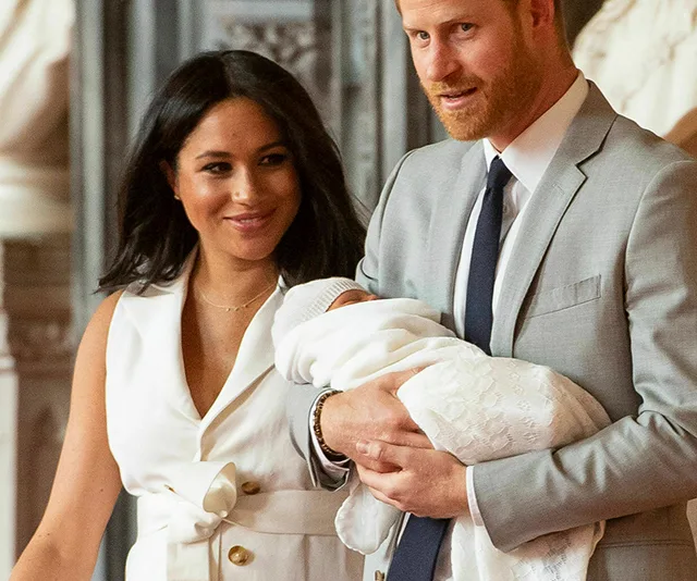 A couple smiling, holding their newborn wrapped in a white blanket after the christening ceremony inside a grand hall.