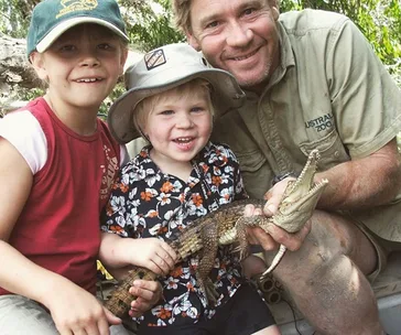 Three people smiling, holding a small crocodile outdoors, with greenery in the background.