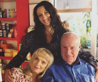 Three people smiling; a woman stands behind an older couple seated, set in a cozy room with shelves in the background.