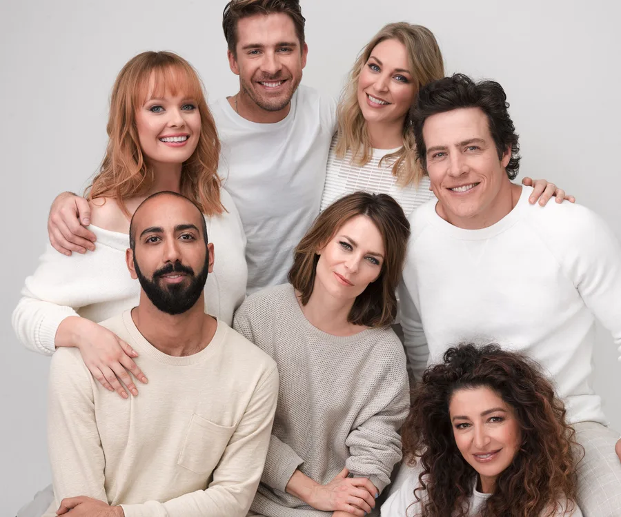 Group photo of seven smiling individuals from a TV series, dressed in white and beige, seated and standing closely together.