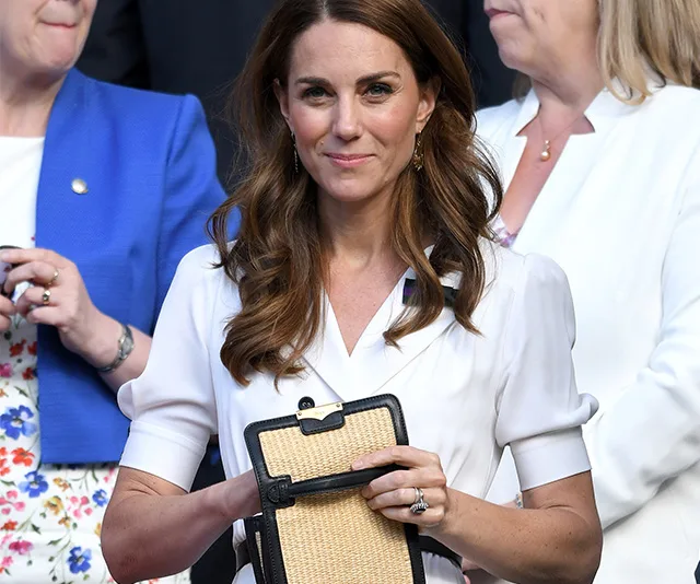 A woman in a white dress holds a straw clutch, standing among others in formal attire.