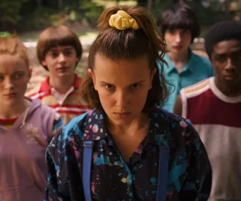 Group of teenagers in 'Stranger Things,' with a girl in front focusing intensely, others stand behind her in casual attire.