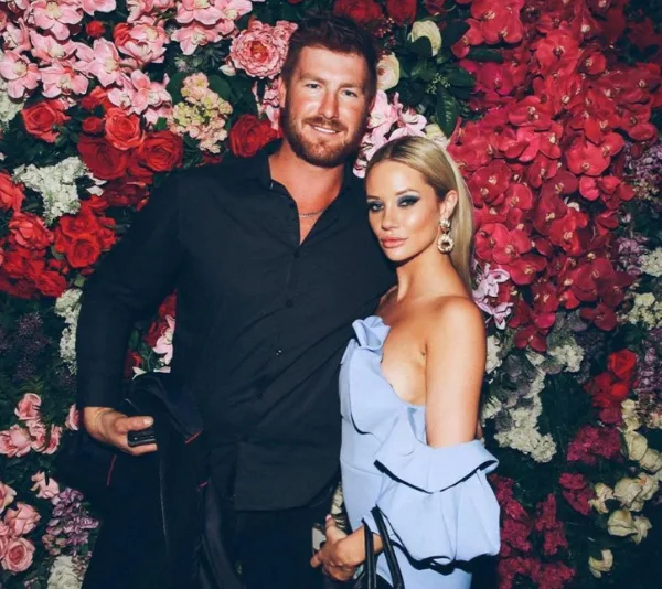 Man and woman pose smiling in front of vibrant floral wall, she wears a light blue dress, he wears a black shirt.