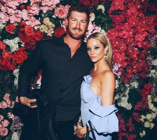Man and woman pose smiling in front of vibrant floral wall, she wears a light blue dress, he wears a black shirt.