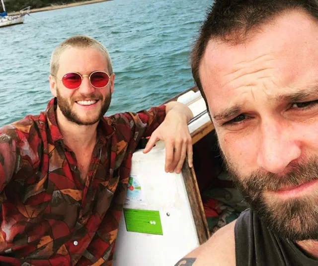 Two men taking a selfie on a boat with the ocean in the background. One wears sunglasses, both have short hair and beards.