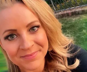 Blonde woman smiling outdoors with green grass and a wrought iron fence in the background.