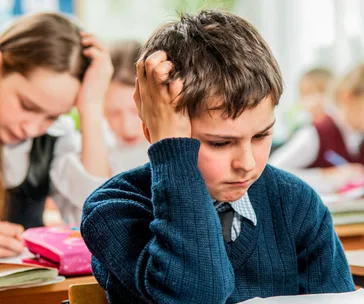 A young boy in a classroom looks frustrated, holding his head while working on an assignment.