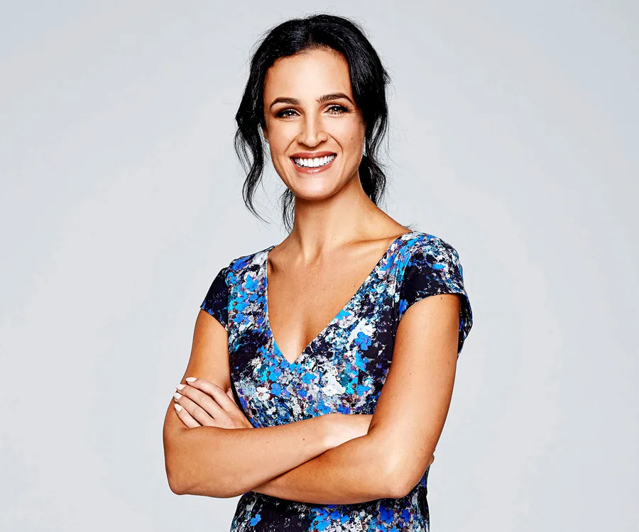 A woman smiling confidently with arms crossed, wearing a blue floral dress against a plain background.