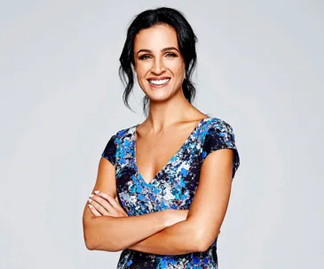 A woman smiling confidently with arms crossed, wearing a blue floral dress against a plain background.