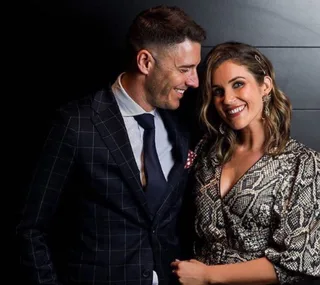 Couple smiling in formal attire, man in a checkered suit, woman in a patterned dress, posing against a dark background.