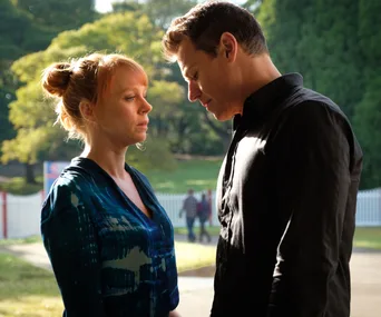 A woman and a man stand close, gazing at each other intensely, with trees and a white fence in the background.