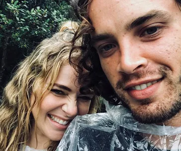 A smiling couple taking a selfie outdoors, wearing rain ponchos, with trees in the background.