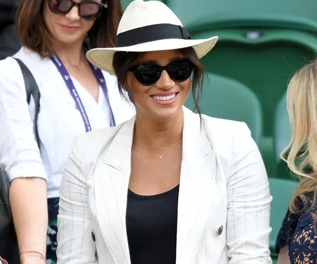 Meghan Markle wearing a white blazer, black top, sunglasses, and a hat, smiling at an event.