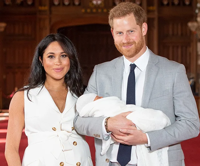 A couple stands together, smiling, inside a formal setting, with one holding a baby wrapped in a white blanket.