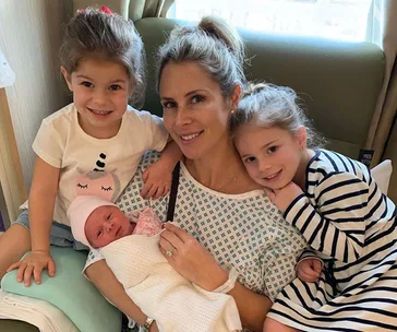 A woman in a hospital gown holds a newborn, surrounded by two smiling young girls.