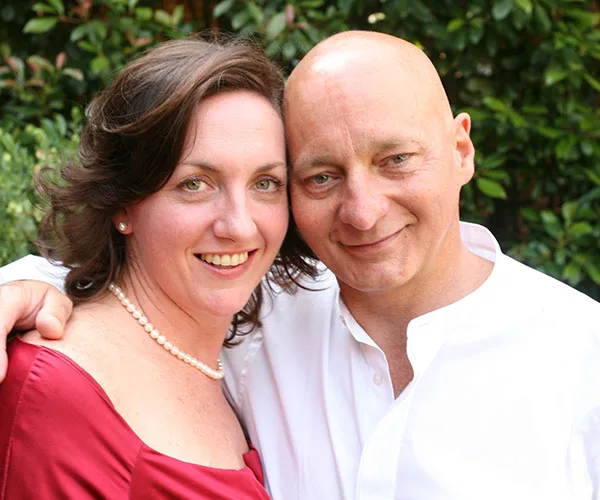 A smiling couple, the woman in a red dress and pearl necklace, the man in a white shirt, posing closely together outdoors.