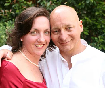 A smiling couple, the woman in a red dress and pearl necklace, the man in a white shirt, posing closely together outdoors.