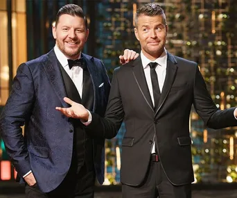 Two men in suits standing on a TV set with a colorful background, one smiling, the other shrugging slightly.