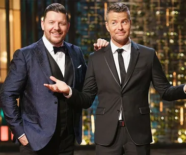 Two men in suits standing on a TV set with a colorful background, one smiling, the other shrugging slightly.
