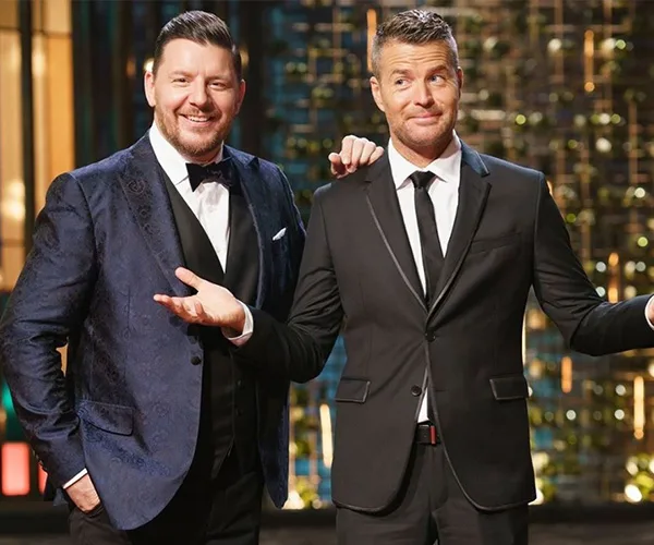 Two men in suits standing on a TV set with a colorful background, one smiling, the other shrugging slightly.