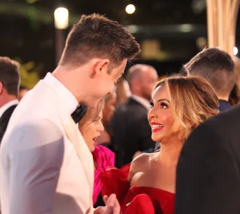 Man in white tuxedo and woman in red dress smiling at each other at a formal event.