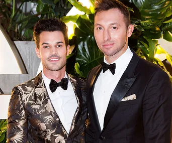 Two men in formal attire, one in a floral jacket, the other in a black suit, posing together at an event.