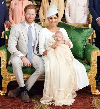 A couple sits on a green couch with a baby in a christening gown, surrounded by elegantly dressed people.