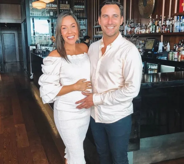 A smiling couple pose together in a bar, with the woman in a white dress touching her belly.