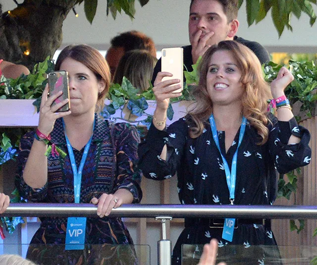Two women with VIP passes enjoying a concert, capturing moments with their phones, surrounded by greenery.