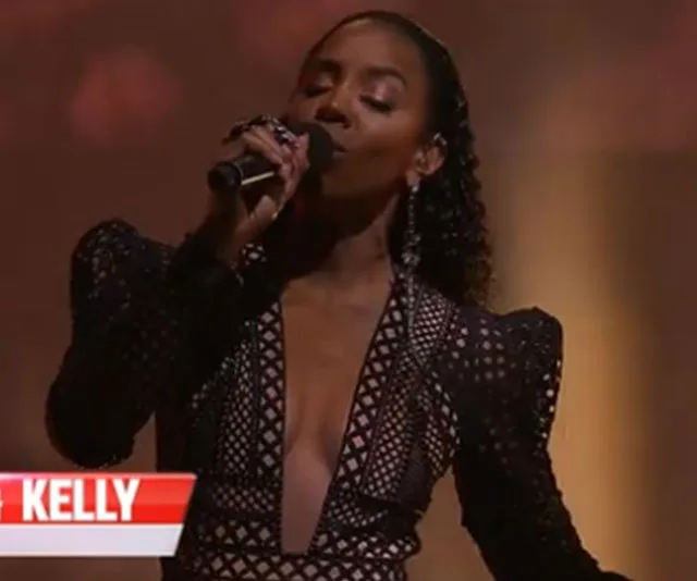 Singer in a black, geometric-patterned dress performs on stage with a microphone, spotlight illuminating her.