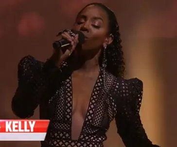 Singer in a black, geometric-patterned dress performs on stage with a microphone, spotlight illuminating her.