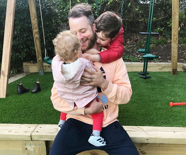 Father hugging two young children in a garden with swings in the background.