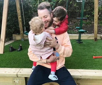 Father hugging two young children in a garden with swings in the background.
