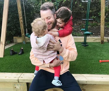 Father hugging two young children in a garden with swings in the background.