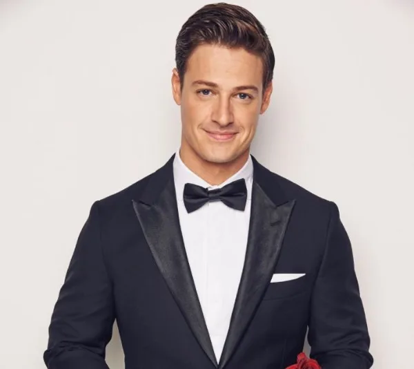 Man in a tuxedo smiling against a plain background.