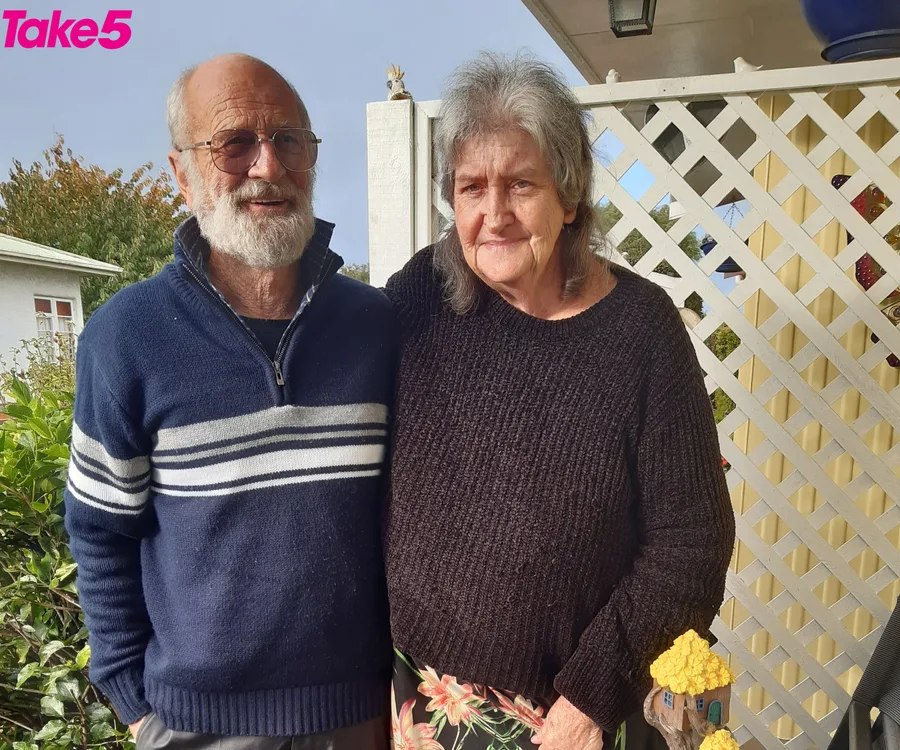 Elderly couple smiling, standing close together outside, with a white lattice and greenery in the background.