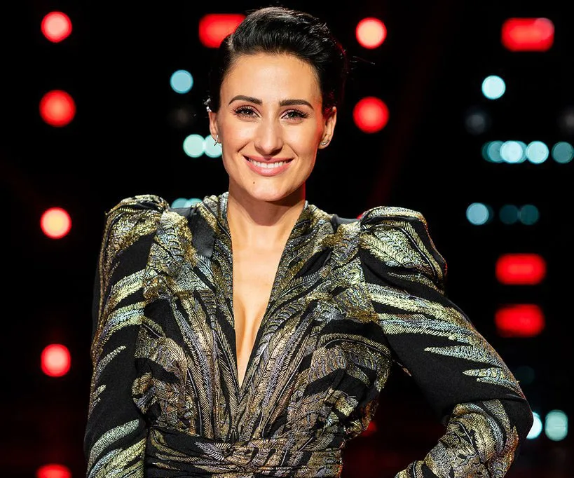 Diana Rouvas smiling in a glamorous outfit on The Voice Australia stage, with colorful lights in the background.