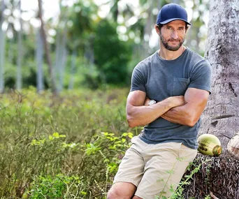 Man in casual clothes and cap leans against a tree outdoors, surrounded by tropical foliage and coconuts.