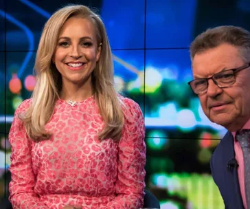 "Two people on a TV set, one in pink dress smiling, the other in glasses and suit."