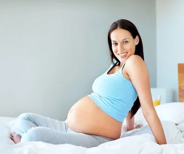 Pregnant woman smiling, sitting on a bed in comfortable clothing.