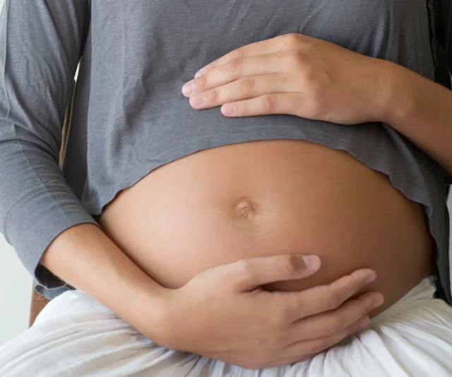 Pregnant woman at 32 weeks holding her belly, wearing a gray top and white skirt.