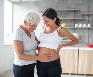 Two women smiling and touching a pregnant belly in a modern kitchen setting.