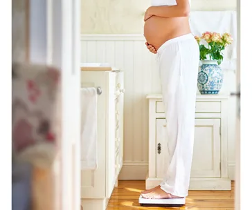 Pregnant woman at 35 weeks standing on a scale in a bathroom, side profile, wearing white clothes.