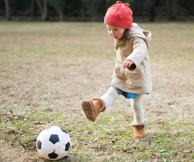 Toddler in a beige coat and red hat kicks a soccer ball on grass.