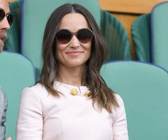A woman in sunglasses wearing a light-colored outfit with a gold necklace, seated outdoors with green seats in the background.