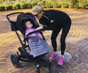 A woman bends down to interact with a baby in a stroller outdoors on a sunny day.