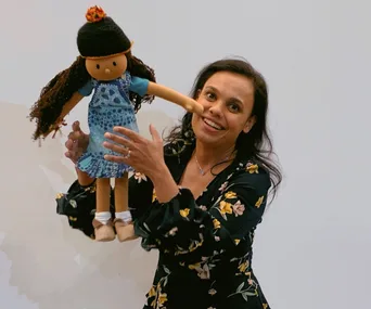 Miranda Tapsell holds the Play School character Kiya, a doll with long hair, wearing a blue dress and black beanie.