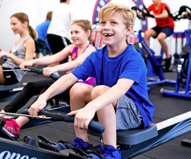 Kids smiling while exercising on rowing machines in a fitness studio.