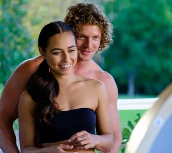 A smiling woman and man, standing close outdoors, with green foliage in the background.